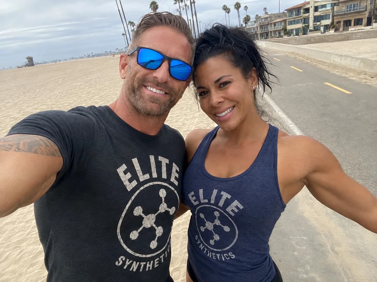Two people wearing Elite Synthetics shirts on a beachfront path.